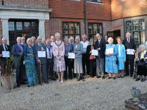 A group of older adults, some holding certificates, pose together outside a brick building. Most are dressed formally and stand on a gravel area, smiling at the camera.
