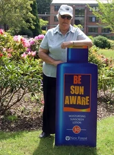 An older adult wearing sunglasses and a cap stands outdoors by a flower bed, leaning on a large sign shaped like a sunscreen bottle that says Be Sun Aware and Moisturising Suncream Lotion SPF 30.