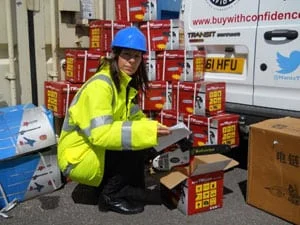 A person wearing a blue hard hat and a bright yellow safety jacket kneels beside stacked red boxes near a white van, holding paperwork or a checklist outdoors.