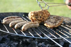 Four sausages and two pieces of seasoned meat are being grilled over an outdoor barbeque, with a pair of tongs holding one piece. The background is blurred green grass.
