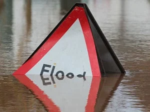 A triangular flood warning sign is partially submerged in water, with only the top visible above the floodwater. The word Flood is written on the sign.