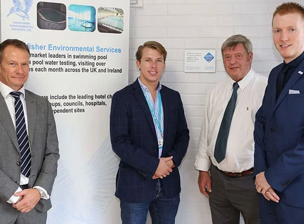Four men in business attire stand indoors, smiling for a photo in front of a white brick wall and a banner for Kingfisher Environmental Services. The banner highlights the company’s swimming pool water testing and services.