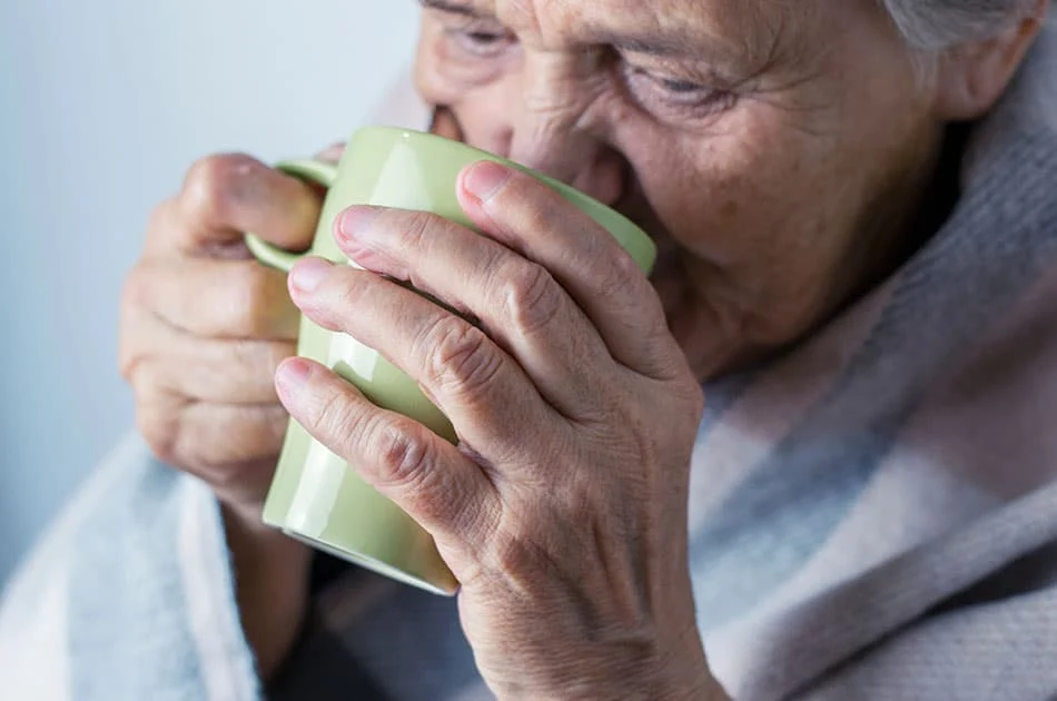 An elderly person wrapped in a blanket holds a green mug with both hands and drinks from it, appearing to enjoy a warm drink.