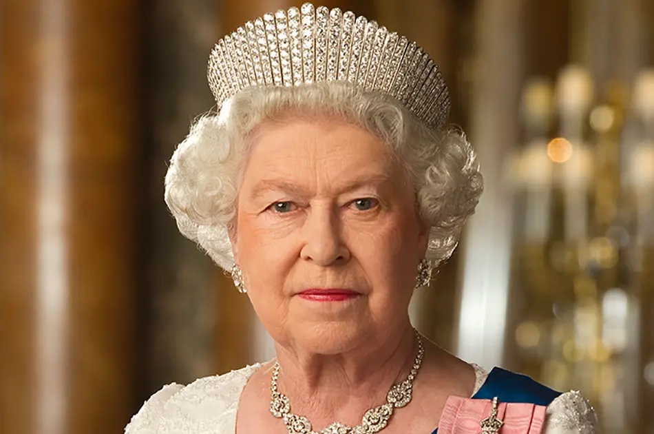 An older woman with white hair, wearing a jewelled tiara, pearl earrings, a necklace, and a white dress, looks directly at the camera against an elegant background.