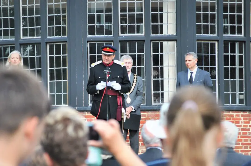 A uniformed official with a red-trimmed hat speaks at a microphone in front of a building, whilst two men in suits and a blurred crowd look on.