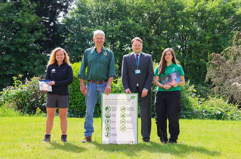Four people stand outdoors on grass with trees in the background, posing beside a sign that displays eco-friendly tips. Three of them hold booklets, and one is dressed in a suit. It is a sunny day.