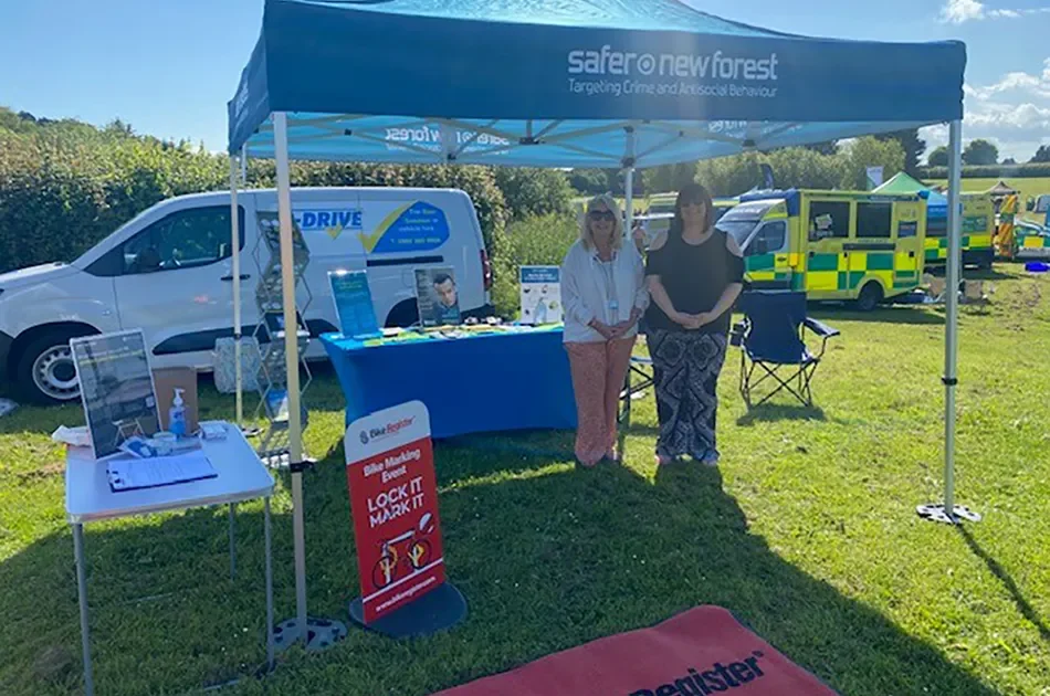 Two women standing under a blue 'Safer New Forest' gazebo at an outdoor community event, with information tables, a van, and emergency vehicles in the background on a sunny day.