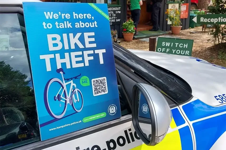 A blue sign reading we're here to talk about bike theft is displayed on a police car. the sign includes an image of a bicycle, a qr code, and a web address. plants and a reception area are visible in the background.