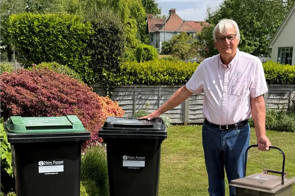 An older man in glasses and a light pink shirt stands in a garden, holding a small brown bin and touching a large black bin. another bin sits nearby. there are bushes, a wooden fence, and houses in the background.