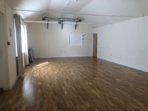 A spacious, empty room with light coloured walls and a wooden floor. there are windows with curtains on the left, a door and white noticeboard on the far wall, and ceiling lighting with exposed beams above.