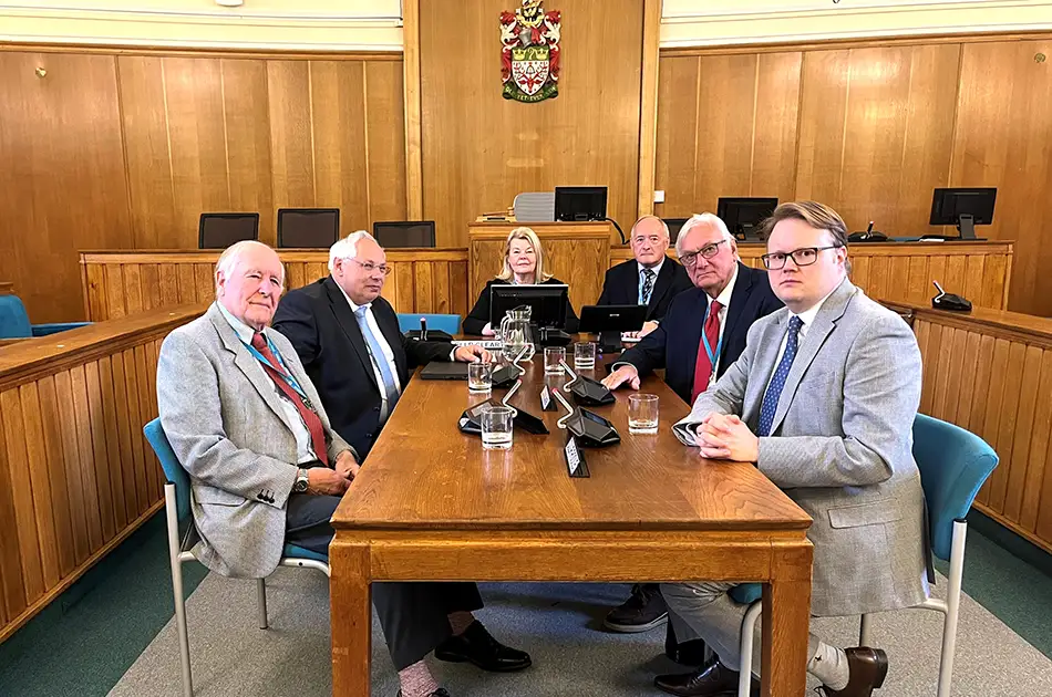 Six people, four men and two women, sit around a wooden table with glasses of water and microphones in a formal courtroom setting with wood panelled walls and a crest behind them.