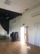 A bright, empty room with a wooden floor, stacked chairs, a green sofa, and an exit door letting in sunlight. black stage curtains and ceiling lights are visible on the left, with white walls and a white curtain on the right.
