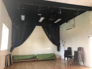 A small stage with black curtains in a bright room. two green covered benches are in front of the stage, and a stack of chairs is nearby. the floor is wooden, and lighting fittings hang from the black ceiling above the stage.