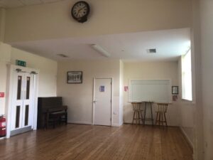 A spacious, empty room with wooden floors, a wall clock, a piano, a small table with two chairs by a window, double doors, a fire extinguisher, and a closed service hatch.