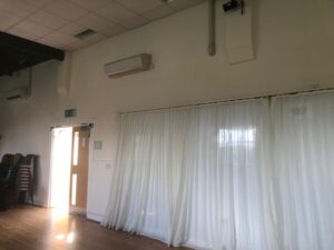A bright room with wooden floors, cream coloured walls, white ceiling tiles, and an air conditioning unit. sheer white curtains cover large windows, and sunlight streams through an open door on the left. stacked chairs are visible in the corner.