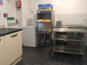 A clean kitchen area with a metal dishwasher, stainless steel trolley, small white freezer, worktop, and shelves. cleaning supplies and trays are on the dishwasher. the floor is dark wood, and the walls are white.