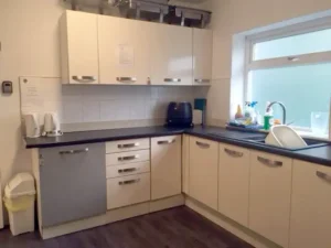A modern kitchen with white cupboards, black worktops, a sink with a draining rack, a window, electric kettle, toaster, bin, and various cleaning supplies on the worktop.