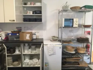 A kitchen area with open cupboards and shelves displaying stacked plates, cups, bowls, a microwave, and a mini fridge. a cardboard box and kitchen items are on the worktop. the space is organised but busy.