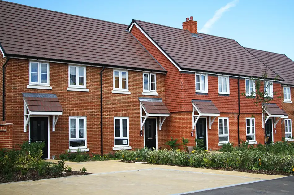 A row of modern, two storey red brick terraced houses with dark brown roofs, white framed windows, small front canopies, and neat landscaping along a clean paved path.