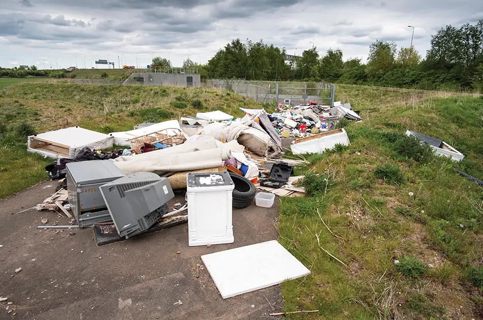 A pile of dumped rubbish, including appliances, tyres, carpets, and debris, lies on a paved area beside grass and a fence under a cloudy sky, with trees and a road in the background.