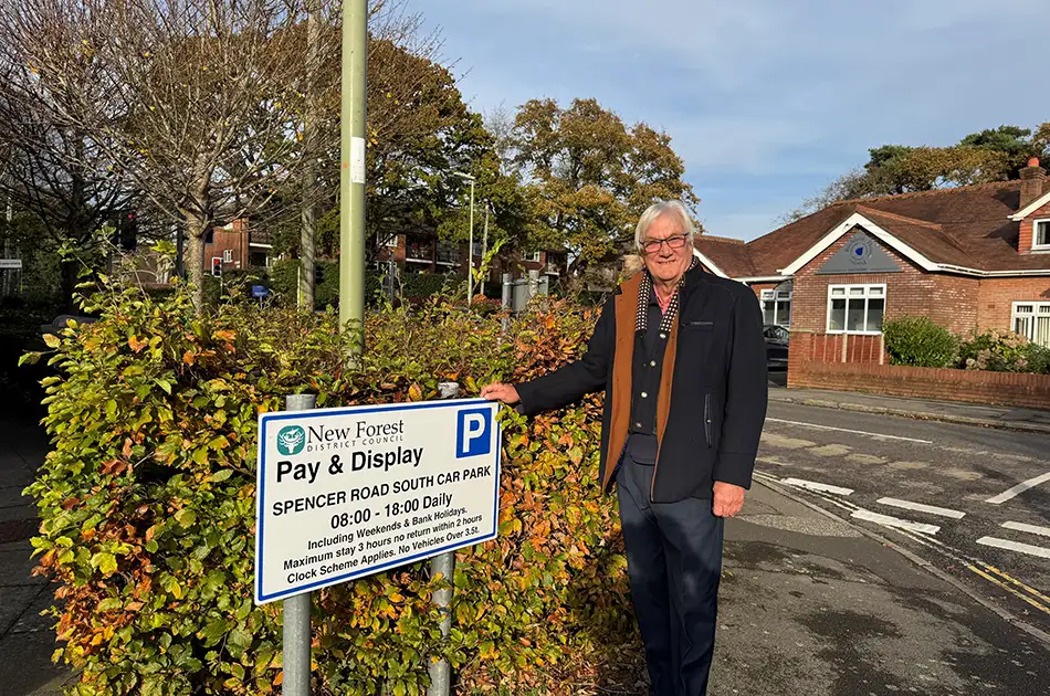 An older man with grey hair and glasses stands by a “Pay & Display” car park sign on Spencer Road South, with houses, greenery, and a sunny blue sky in the background.