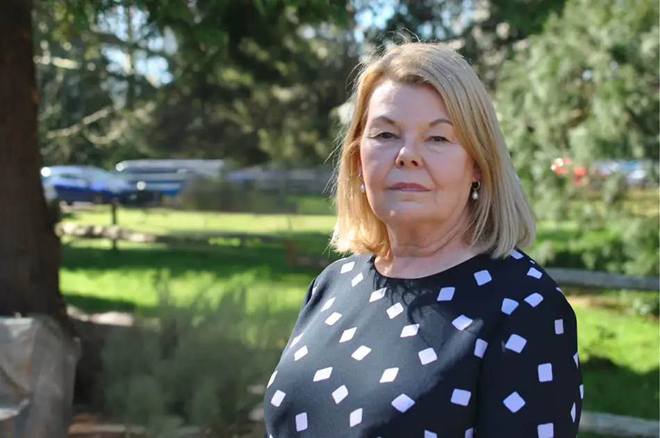 A woman with shoulder length blonde hair stands outdoors in a sunlit park, wearing a dark top with white square patterns. Trees and grass are visible in the background.
