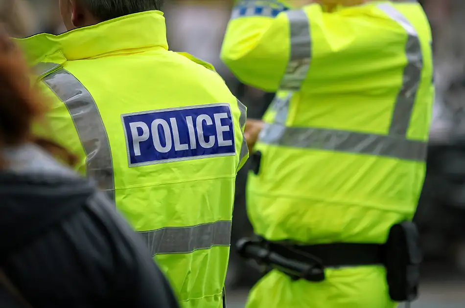 Two people wearing bright yellow jackets labelled POLICE with reflective strips, seen from behind, standing outdoors.