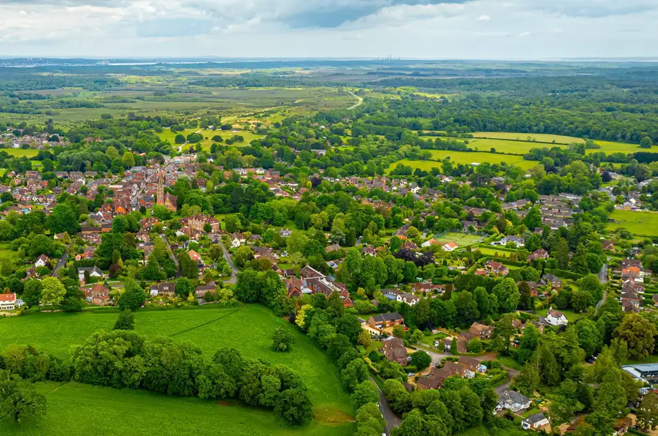 Aerial view of a small village surrounded by lush green fields and dense trees, with houses scattered throughout and expansive countryside stretching into the distance under a cloudy sky.