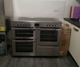 A modern stainless steel cooker with four glass doors and a hob, situated in a kitchen between a worktop and a large cardboard box on a dark wooden floor.