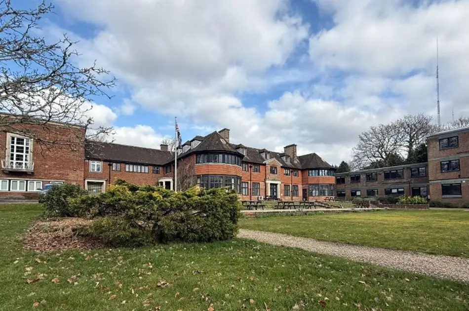 A large brick building with multiple wings and chimneys stands behind a well-kept lawn and hedges under a partly cloudy sky. Paths and outdoor seating are visible in the garden area.