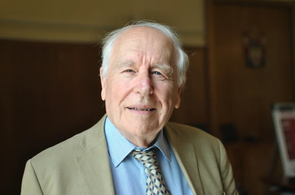 An elderly man with white hair, wearing a beige suit jacket, blue striped shirt, and patterned tie, smiles softly whilst standing indoors in a warmly lit room with wooden walls.