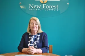 A woman sits at a table with her hands folded, in front of a teal wall displaying a New Forest District Council sign. She is wearing a navy blue blazer over a patterned blouse.