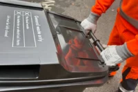 A person wearing orange work clothes and grey gloves is wheeling a black general waste bin with a closed lid on a paved surface.