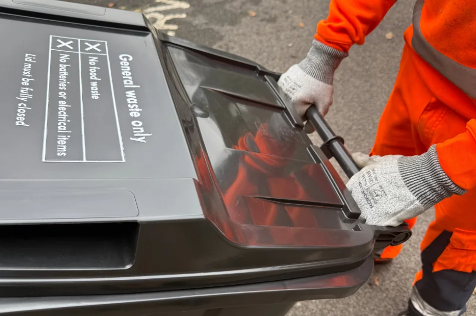 A person wearing orange work clothes and grey gloves is wheeling a black general waste bin with a closed lid on a paved surface.