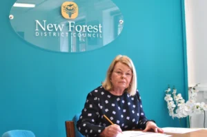 A woman with blonde hair sits at a desk, writing with a pencil, in front of a teal wall with a “New Forest District Council” sign. White flowers and documents are on the desk.