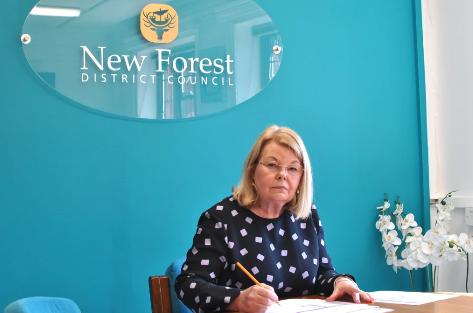 A woman with blonde hair sits at a desk, writing with a pencil, in front of a teal wall with a “New Forest District Council” sign. White flowers and documents are on the desk.