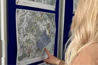 A person with long blond hair points at a detailed map on a wall, which shows various markings and highlighted areas.