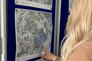 A person with long blond hair points at a detailed map on a wall, which shows various markings and highlighted areas.